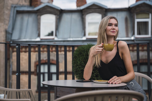 Porträt einer blonden jungen Frau, die auf einer Dachterrasse sitzt und eine Tasse Kaffee trinkt - KKAF02629