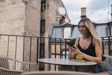 Lächelnde blonde Frau, die mit einer Tasse Kaffee auf der Dachterrasse sitzt und ihr Mobiltelefon überprüft - KKAF02634