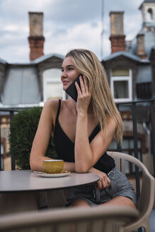 Lächelnde blonde Frau am Telefon auf der Dachterrasse sitzend mit einer Tasse Kaffee - KKAF02635