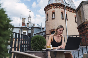 Junge Frau sitzt auf einer Dachterrasse und benutzt einen Laptop - KKAF02636