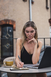 Portrait of young woman sitting on balcony with laptop taking notes - KKAF02639