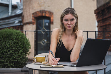 Portrait of young woman sitting on balcony with laptop taking notes - KKAF02640