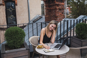Lächelnde junge Frau sitzt mit Laptop auf dem Balkon und macht sich Notizen - KKAF02643