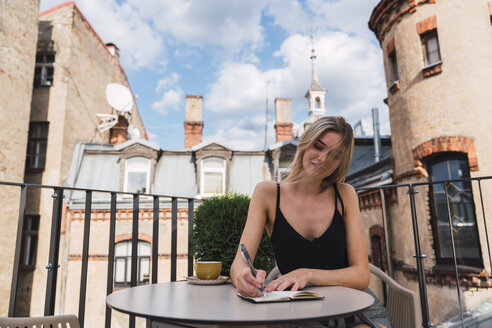 Junge Frau sitzt mit einer Tasse Kaffee auf dem Balkon und macht sich Notizen - KKAF02647