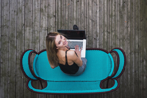 Lächelnde junge Frau sitzt auf einer türkisfarbenen Couch auf der Terrasse und benutzt einen Laptop, Ansicht von oben - KKAF02653
