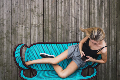 Junge Frau sitzt auf einer türkisfarbenen Couch auf der Terrasse und benutzt ein Smartphone, Ansicht von oben - KKAF02657
