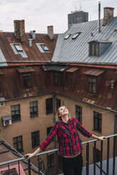 Lächelnde junge Frau steht auf einem Balkon und schaut nach oben - KKAF02666