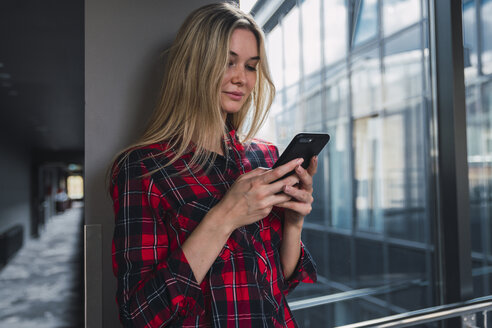 Junge Frau in modernem Bürogebäude schaut auf ihr Handy - KKAF02670