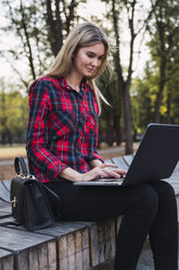 Modische junge Frau sitzt auf einer Bank im Freien und arbeitet an einem Laptop - KKAF02676
