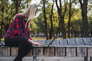 Young woman sitting on bench outdoors working on laptop - KKAF02677