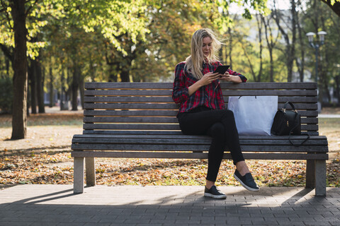 Young woman with shopping bag sitting on bench in autumn using mini tablet stock photo