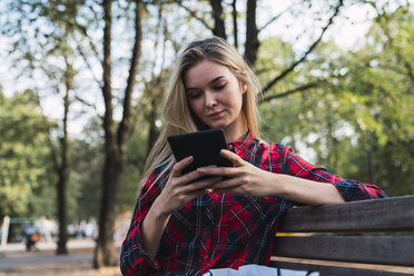 Young woman sitting on bench outdoors using mini tablet - KKAF02687