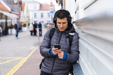 Man leaning against house wall with cell phone and headphones - KKAF02715