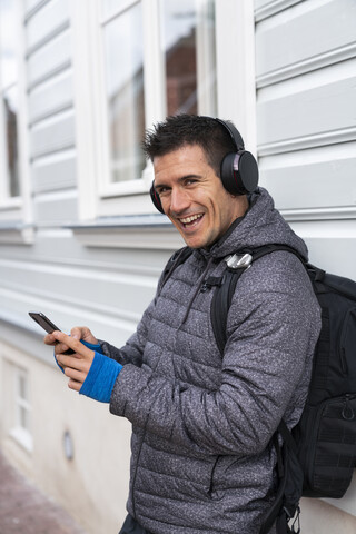 Porträt eines lachenden Mannes an einer Hauswand mit Mobiltelefon und Kopfhörern, lizenzfreies Stockfoto