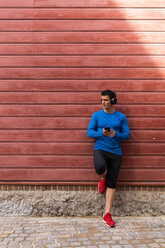 Athlete leaning against house wall holding cell phone - KKAF02718
