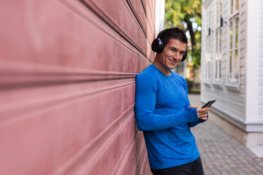 Porträt eines lächelnden Sportlers, der mit Handy und Kopfhörern an einer Hauswand lehnt - KKAF02721