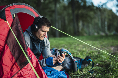 Mann zeltet in Estland, sitzt in seinem Zelt und hört Musik von seinem Smartphone, lizenzfreies Stockfoto