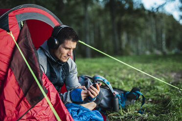 Man camping in Estonia, sitting in his tent, lietsning music from his smartphone - KKA02762