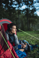 Man camping in Estonia, sitting in his tent, lietsning music from his smartphone - KKA02763