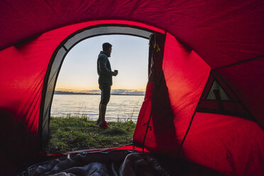 Mann zeltet in Estland, steht vor seinem Zelt und beobachtet den Sonnenuntergang - KKA02796