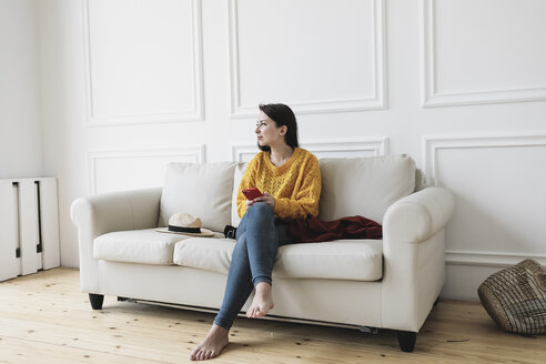 Entspannte junge Frau mit Smartphone sitzt auf der Couch in ihrem neuen Zuhause und schaut in die Ferne - KMKF00595