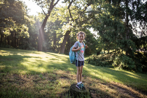 Portrait of girl with backpack standing on tree stump in forest stock photo