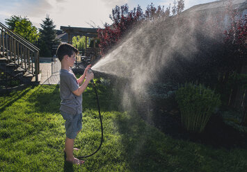 Seitenansicht eines Jungen, der mit einem Gartenschlauch Pflanzen im Garten gießt - CAVF50602