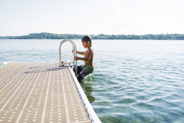 Shirtless boy moving up on ladder in lake against sky - CAVF50632