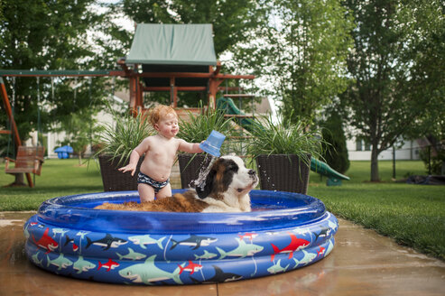 Hemdloser kleiner Junge gießt Wasser auf den Hund, während er im Planschbecken im Garten steht - CAVF50639