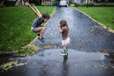 Seitenansicht von verspielten Geschwistern, die sich auf der nassen Straße während der Regenzeit vergnügen - CAVF50657