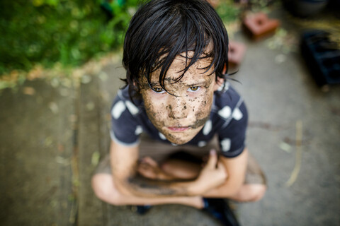 High angle portrait boy with dirty face sitting on footpath at yard during rainy season stock photo