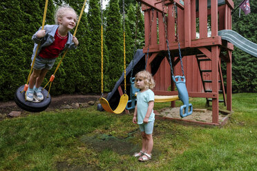 Sad girl looking at happy sister swinging on tire swing at playground - CAVF50666