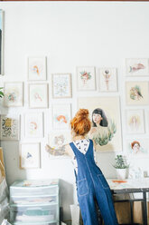 Rear view of female artist hanging paintings on wall in studio - CAVF50679