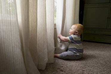 Seitenansicht eines kleinen Jungen, der mit Vorhängen spielt, während er auf dem Teppich zu Hause sitzt - CAVF50730