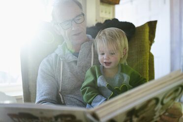 Niedriger Blickwinkel auf Großvater mit Enkelin, die auf einem Stuhl sitzend zu Hause ein Buch liest - CAVF50752