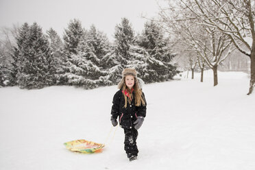 Full length of girl wearing warm clothing while walking with sled on snowy field against trees during snowfall - CAVF50761