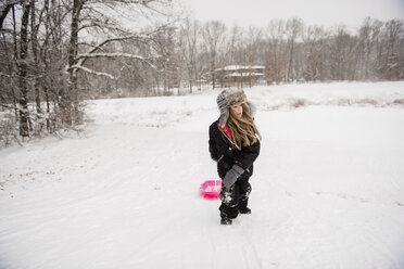 Girl wearing warm clothing while walking with sled on snowy field against trees during snowfall - CAVF50762