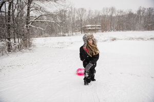 Mädchen trägt warme Kleidung, während sie mit einem Schlitten auf einem verschneiten Feld gegen Bäume läuft - CAVF50762