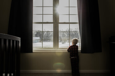 Rear view of boy looking through window while standing at home during winter - CAVF50764