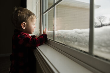 Side view of boy looking through window while standing at home during winter - CAVF50766