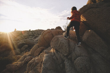 Rückansicht eines Jungen, der bei Sonnenuntergang im Joshua Tree National Park auf Felsen klettert - CAVF50780