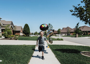 Fröhlicher Junge mit Luftballons, der auf einem Fußweg gegen einen klaren blauen Himmel an einem sonnigen Tag läuft - CAVF50865