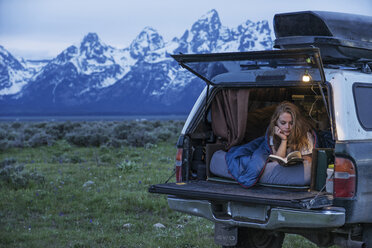 Young woman reading book while relaxing in motor home at forest - CAVF50892