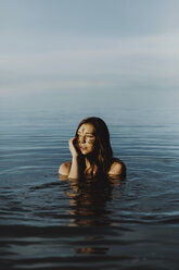 Young woman with mud on face looking away while swimming in lake against sky - CAVF50991