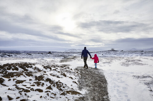 Rückansicht von Vater und Tochter, die sich an den Händen halten, während sie durch eine verschneite Landschaft mit bewölktem Himmel laufen - CAVF51006