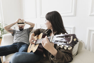 Relaxed couple sitting on couch, woman playing the guitar at home - KMKF00606