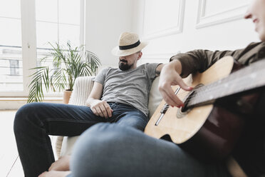 Relaxed couple sitting on couch, woman playing the guitar at home - KMKF00607