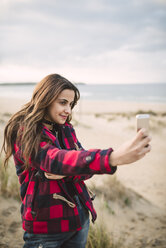 Porträt einer lächelnden jungen Frau, die ein Selfie mit ihrem Smartphone am Strand macht - RAEF02186