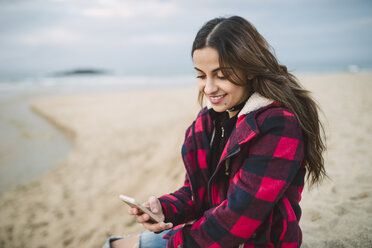 Lächelnde junge Frau mit Smartphone am Strand - RAEF02196