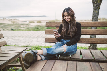 Lächelnde junge Frau sitzt auf einer Bank und schaut auf ihr Handy - RAEF02207